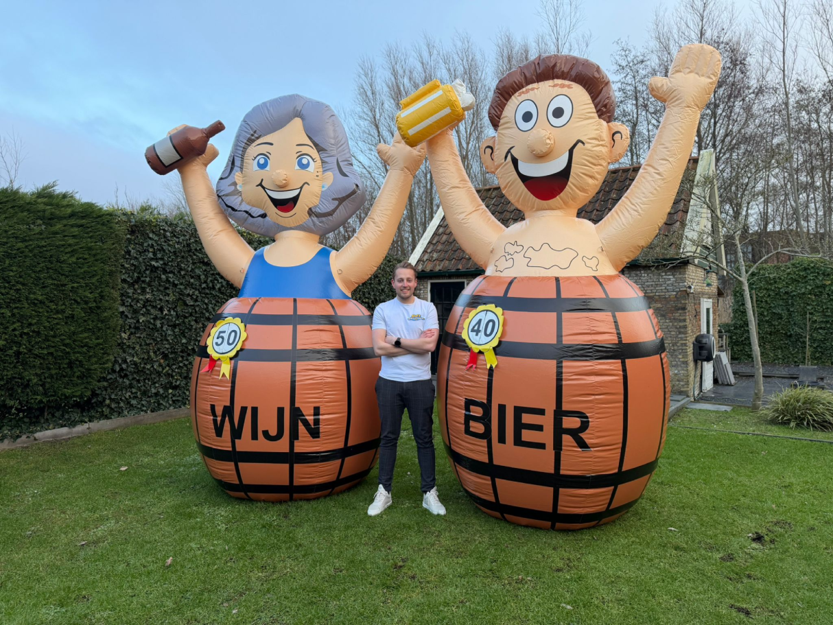 Twee opblaasbare figuren met de teksten "Wijn" en "Bier", staan naast een persoon in de tuin.
